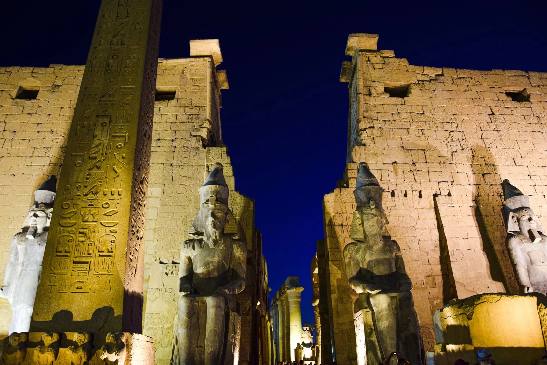 Beleuchteter Tempeleingang mit Obelisk und sitzenden Pharaonenstatuen bei Nacht