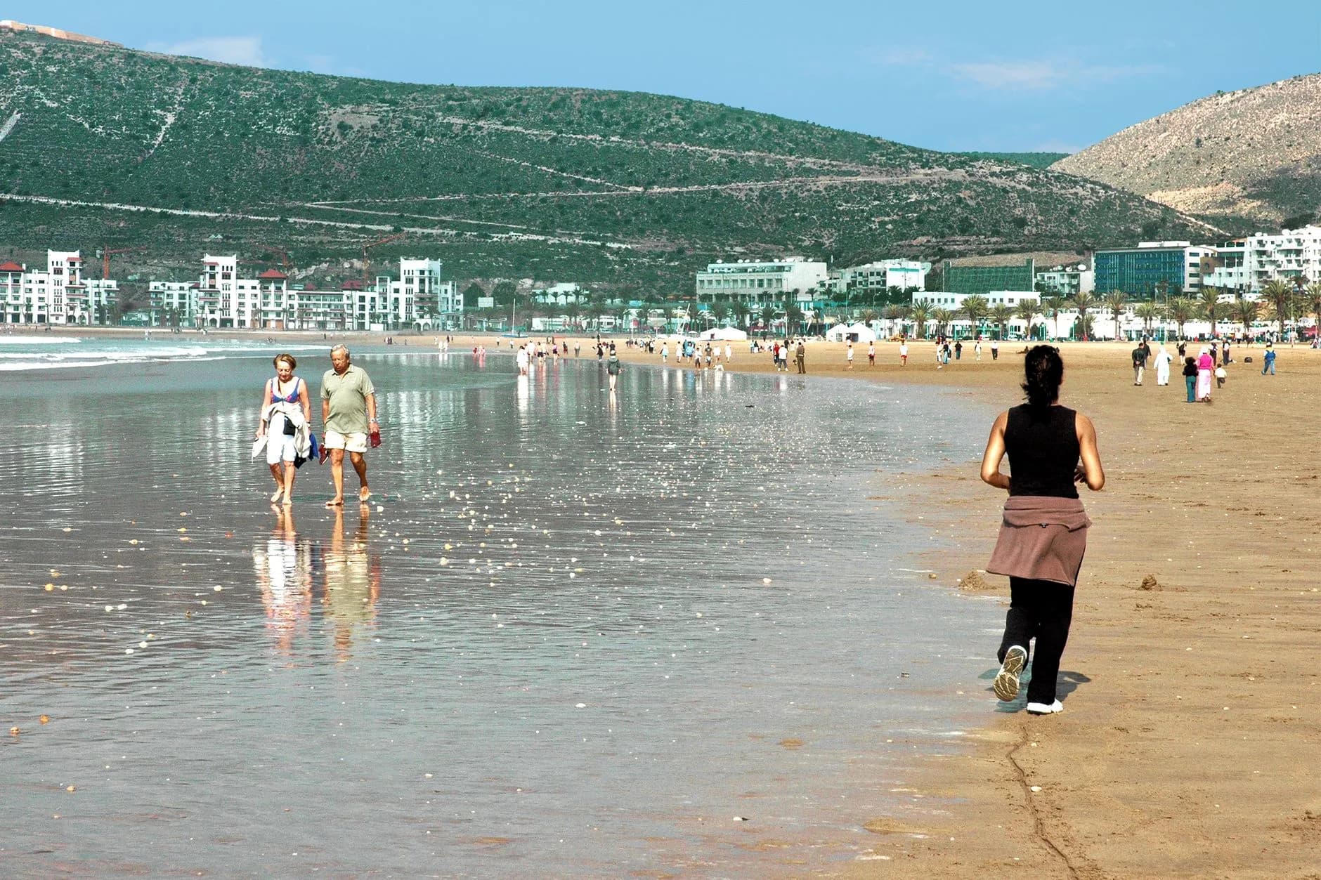 Spaziergänger und Joggerin am flachen Sandstrand vor Küstenstadt mit grünem Berg im Hintergrund