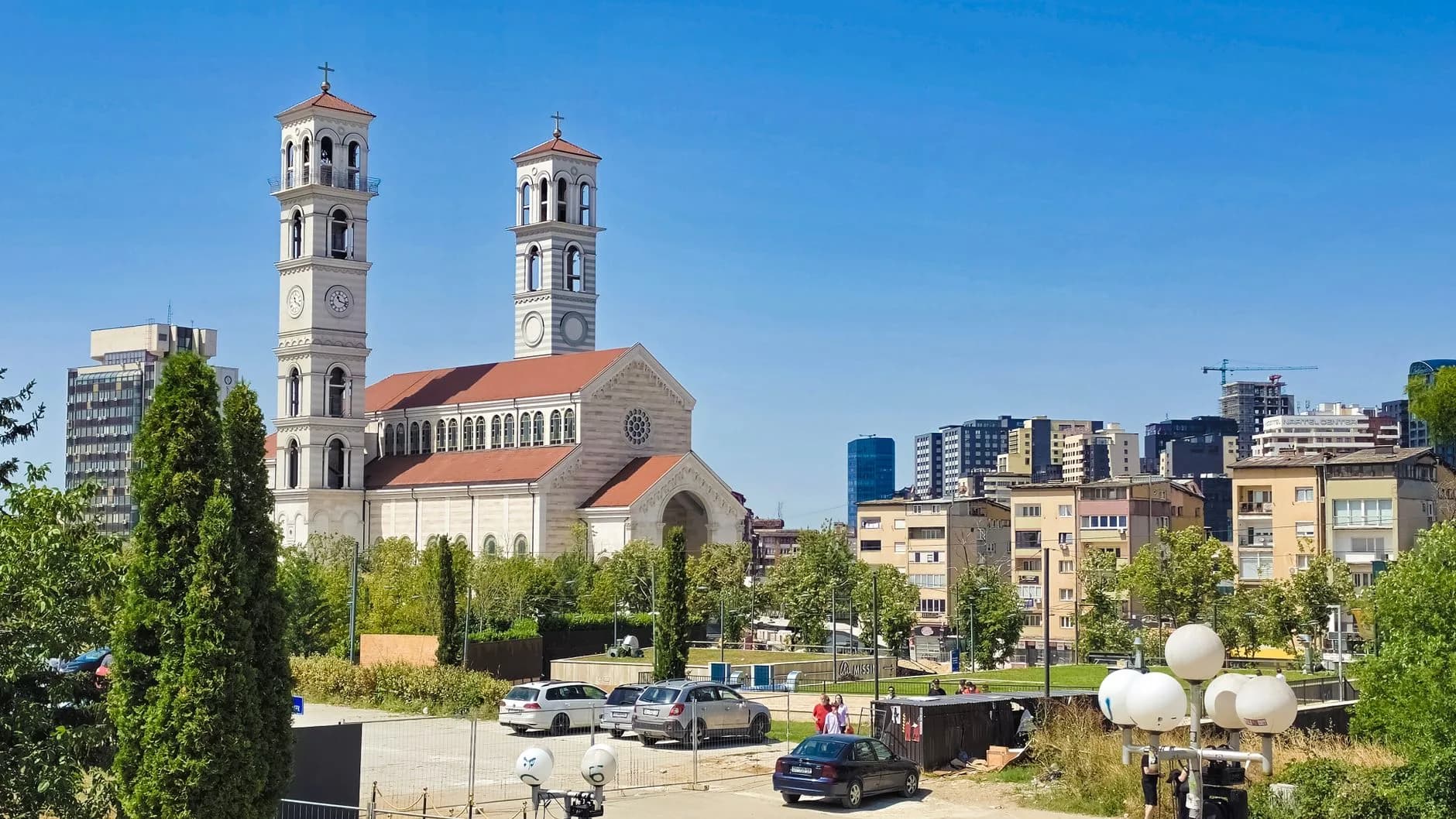 Kirche mit zwei Türmen vor moderner Stadtsilhouette, umgeben von Bäumen und Wohnhäusern
