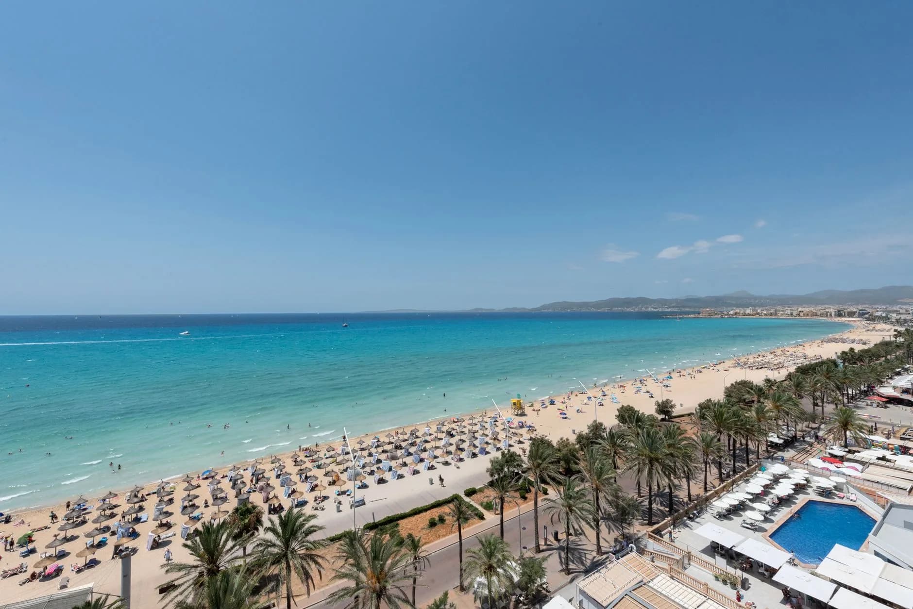Luftaufnahme einer weiten Bucht mit türkisblauem Meer, Sandstrand mit Sonnenschirmen und Palmen an der Promenade