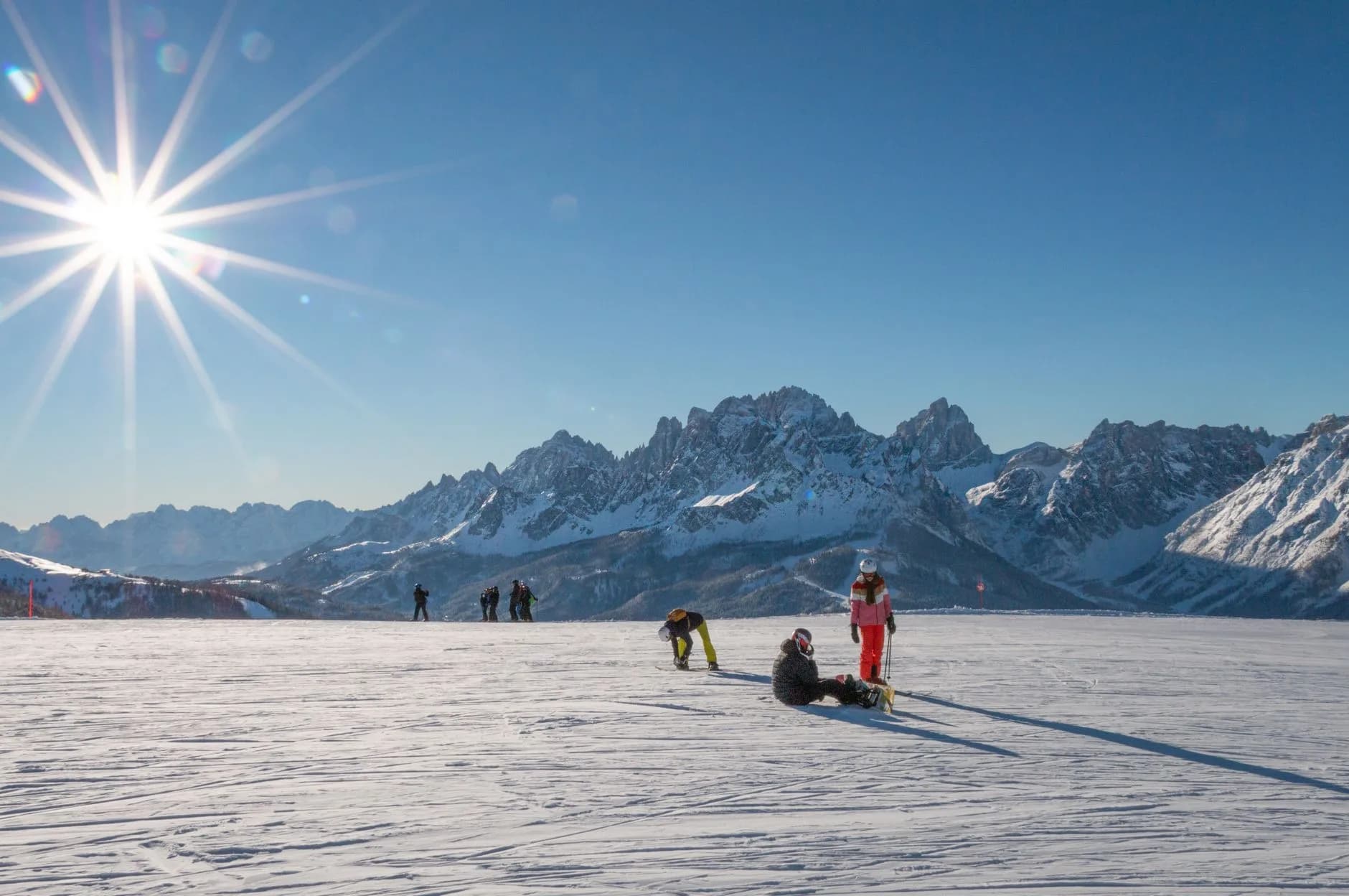 Skifahrer und Snowboarder auf einer sonnigen Piste vor schneebedeckten Berggipfeln unter strahlend blauem Himmel