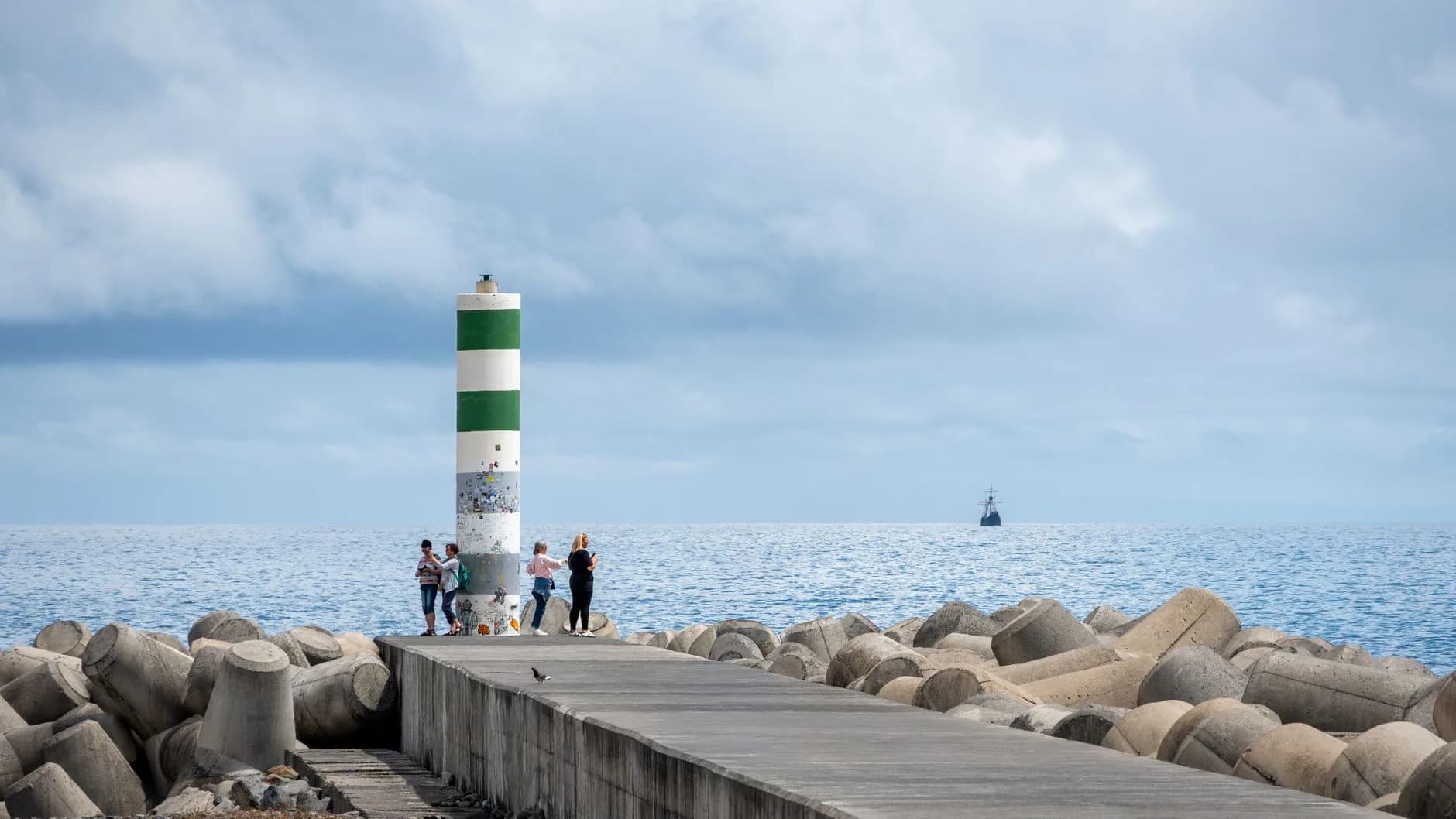 Menschen stehen an einer grün-weißen Hafenbake auf einer Mole aus Wellenbrechern, im Hintergrund ein Segelschiff auf dem Meer