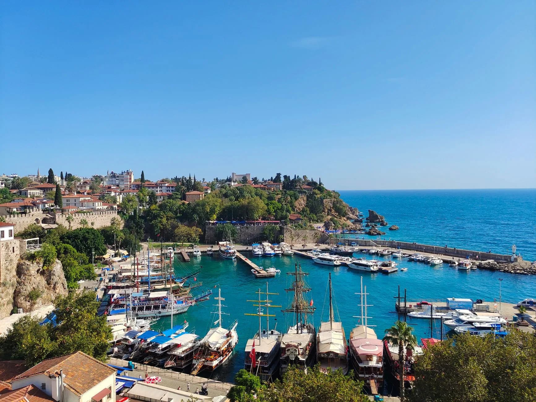Blick von oben auf den alten Hafen von Antalya mit Ausflugsbooten in türkisfarbenem Wasser, umgeben von Altstadt und Küste
