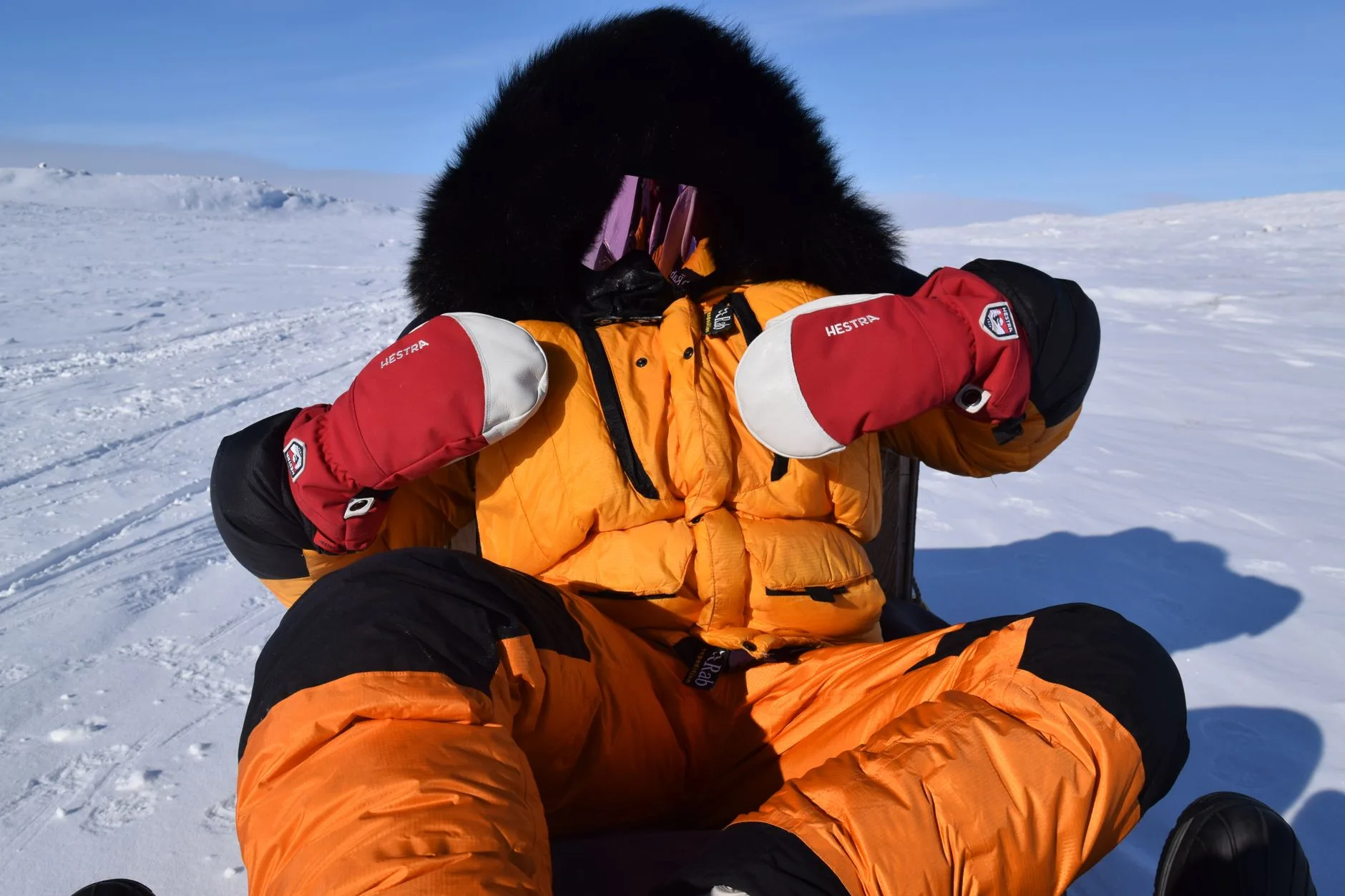 Person in orangefarbenem Expeditionsanzug mit Pelzkapuze und roten Fäustlingen sitzt auf Schnee
