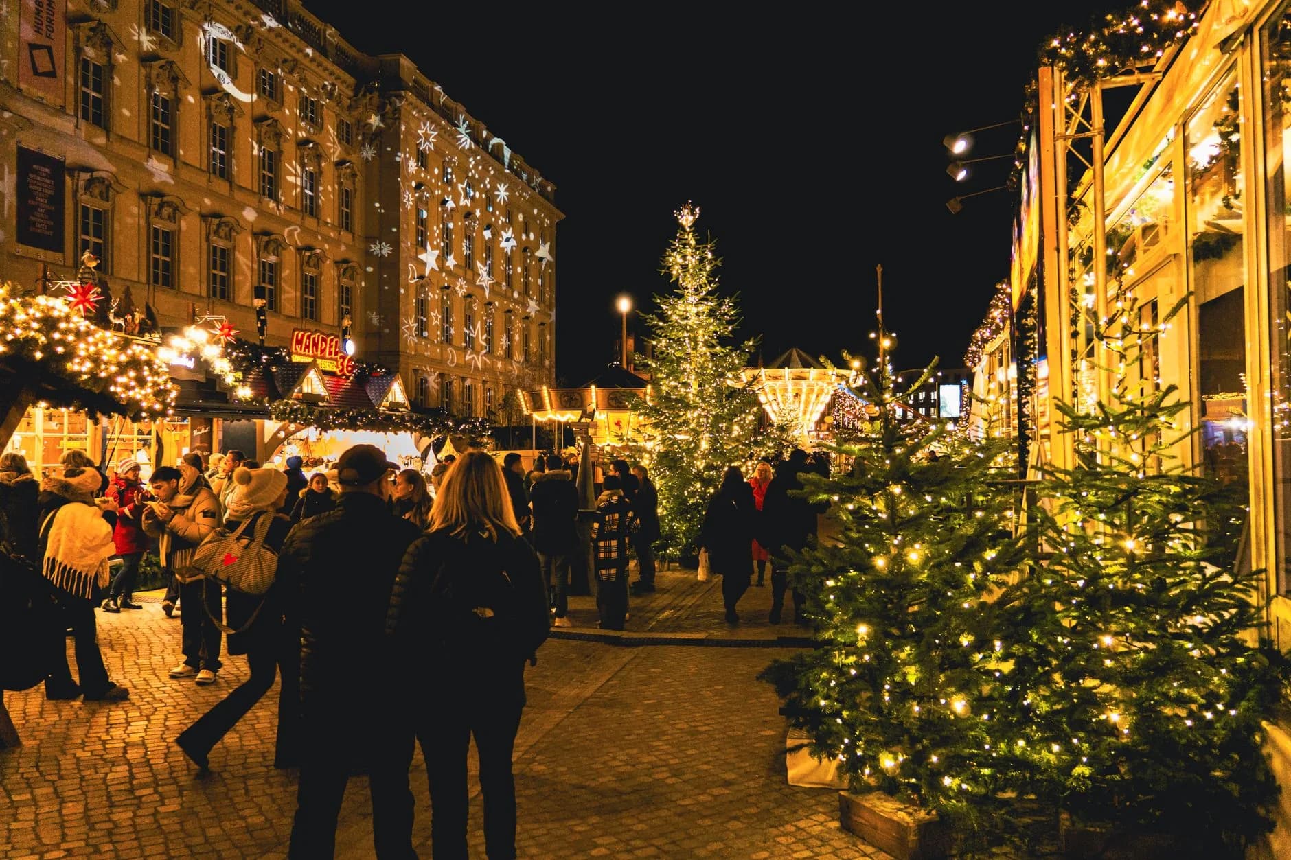 Beleuchteter Weihnachtsmarkt bei Nacht mit geschmückten Tannenbäumen und Besuchern zwischen den Ständen