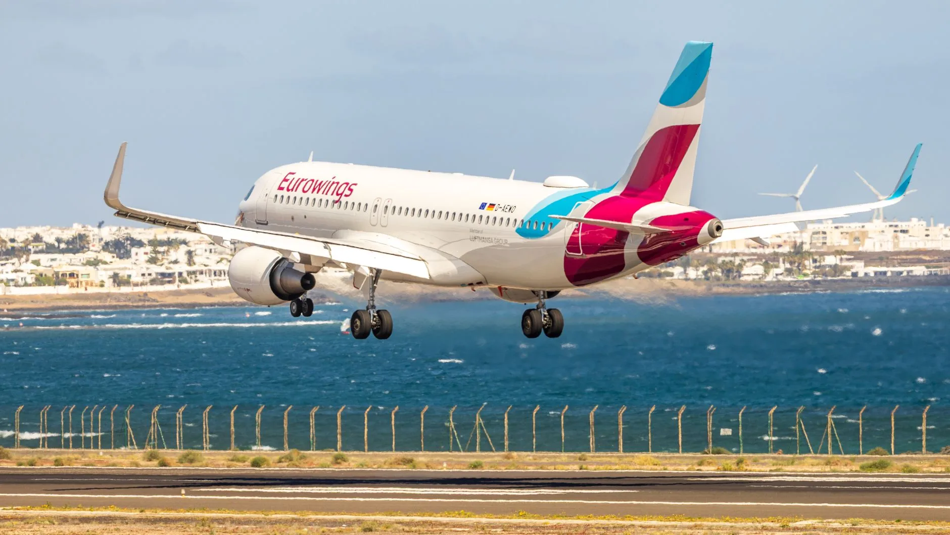 Eurowings-Flugzeug landet im Anflug über einem Zaun nahe der Küste, im Hintergrund Meer und Küstenort