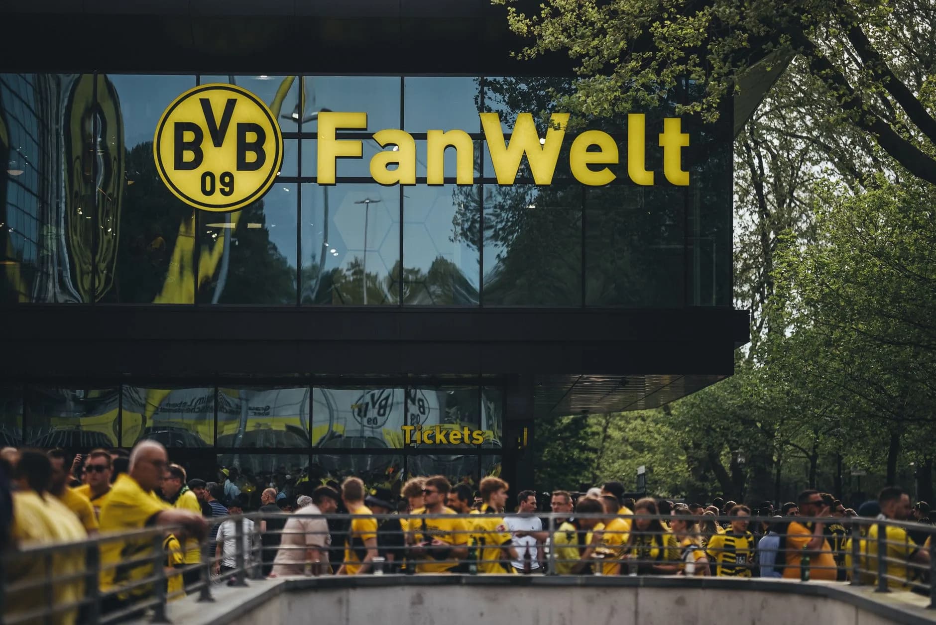 Menschengruppe in gelb-schwarzen Trikots vor der BVB FanWelt mit Glasfassade und Bäumen