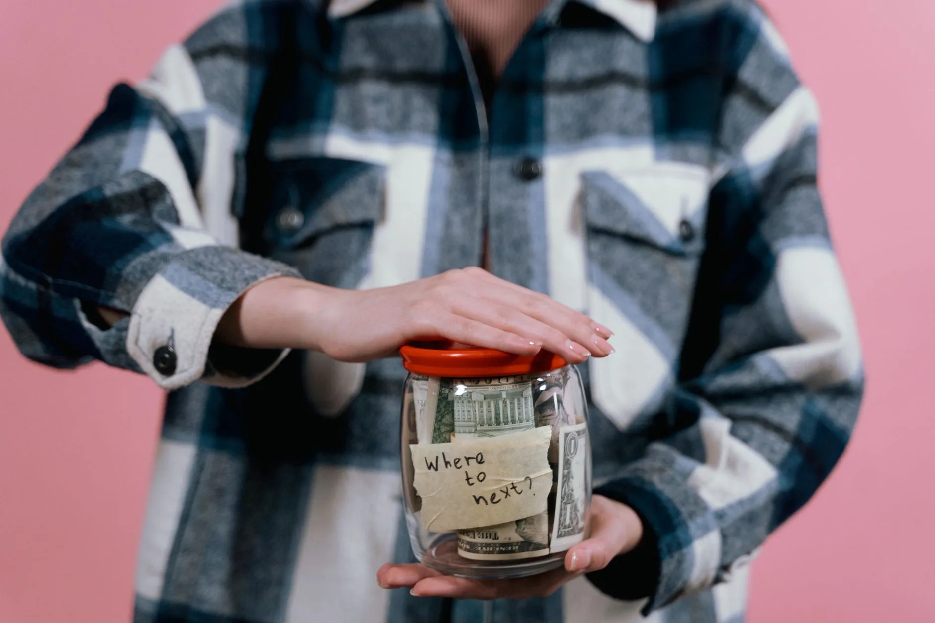 Person im Flanellhemd hält ein Glas mit Dollarscheinen und der Aufschrift 'Where to next?'