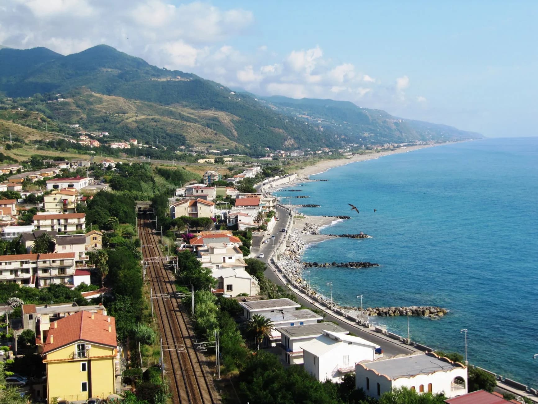 Luftaufnahme einer Küstenstraße mit Bahngleisen zwischen Häusern und dem türkisblauen Meer, im Hintergrund bewaldete Berge