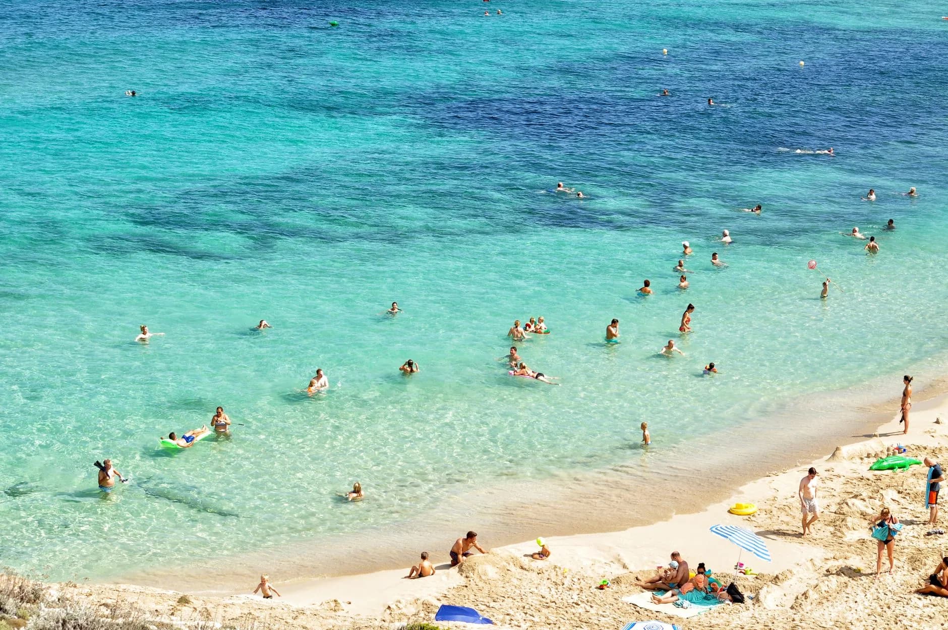Blick von oben auf eine Bucht mit türkisfarbenem Wasser, badende Menschen und Sandstrand mit Sonnenschirmen
