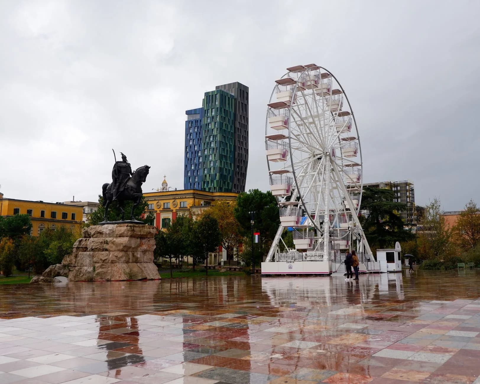 Reiterstatue und Riesenrad auf regennassem Platz vor moderner Skyline und historischen Gebäuden