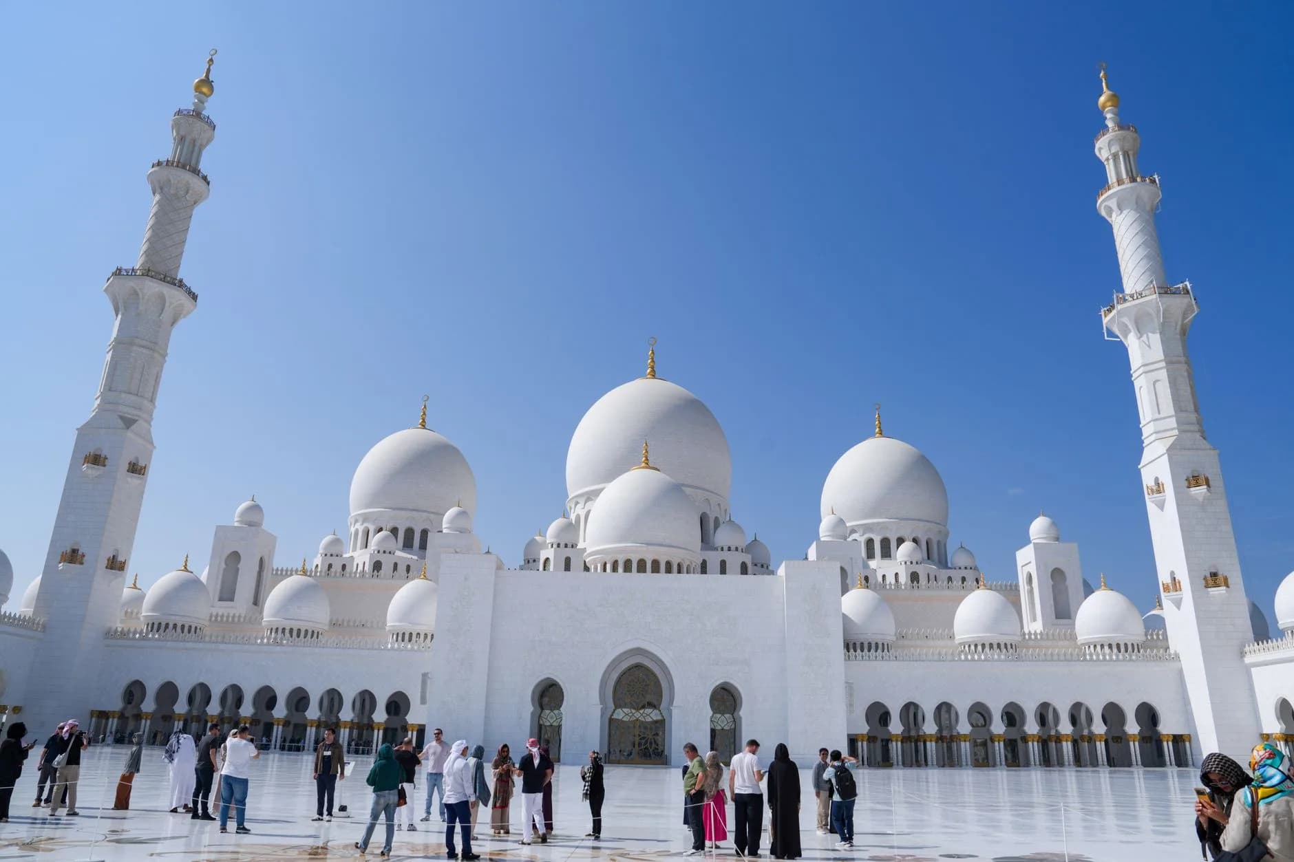 Weiße Moschee mit Kuppeln und Minaretten, Besucher auf dem Marmorvorplatz bei klarem Himmel
