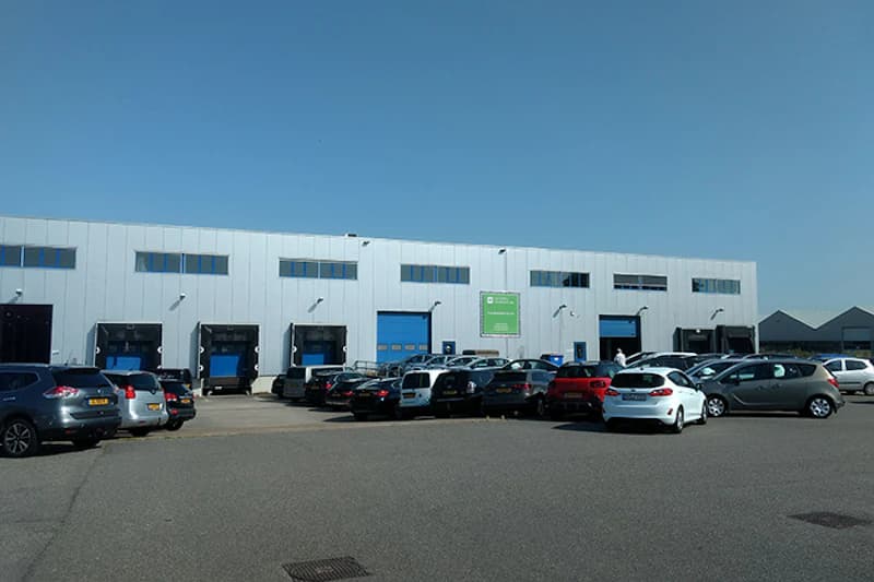 Parkende Autos vor einer Industriehalle mit Ladebuchten und blauen Toren unter klarem Himmel.