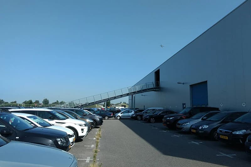 Reihen geparkter Autos neben einer grauen Industriehalle mit erhöhter Rampe und Flugzeug am Himmel.