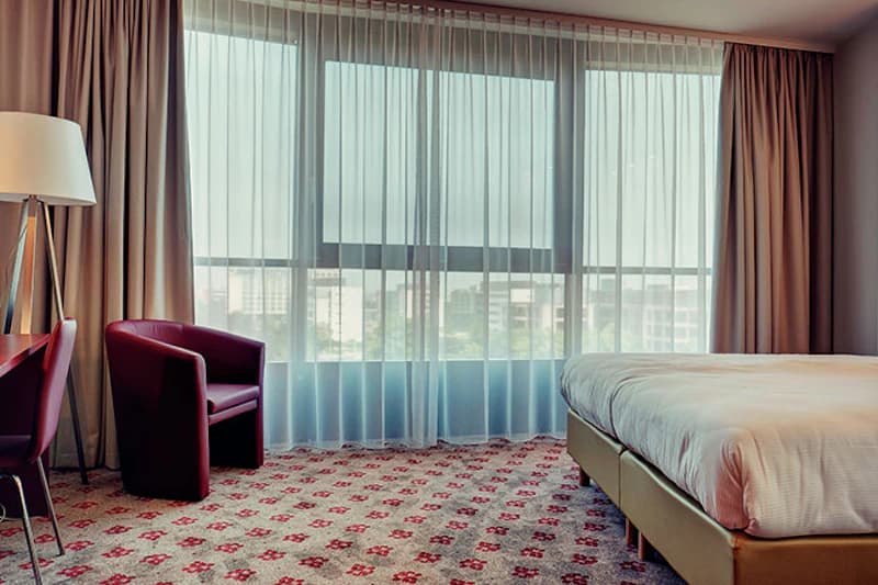 Hotelzimmer mit großem Fenster, halbtransparenten Vorhängen, Doppelbett, rotem Sessel, Stehlampe und gemustertem Teppich.