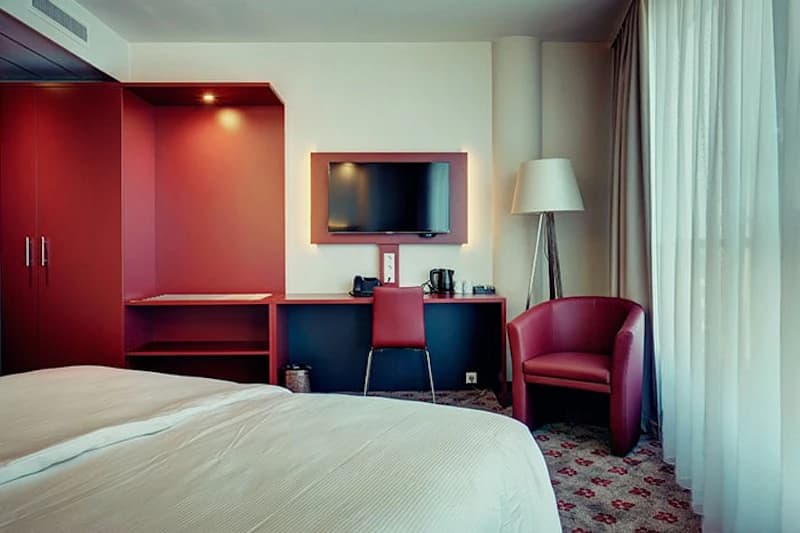 Hotelzimmer mit Bett, rotem Schreibtisch und Einbauschrank, Wandfernseher, Sessel und Fenster mit Gardinen.