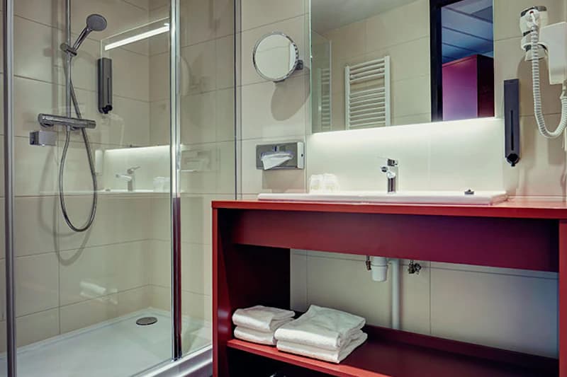 Hotelbadezimmer mit Glasdusche, Duschkopf, rotem Waschtisch, Handtüchern und beleuchtetem Spiegel.