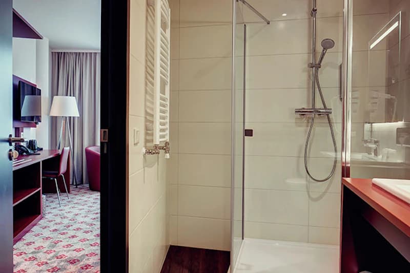 Badezimmer mit Glastürdusche, Handbrause, Handtuchheizkörper und Blick ins angrenzende Zimmer mit Schreibtisch und Fernseher.
