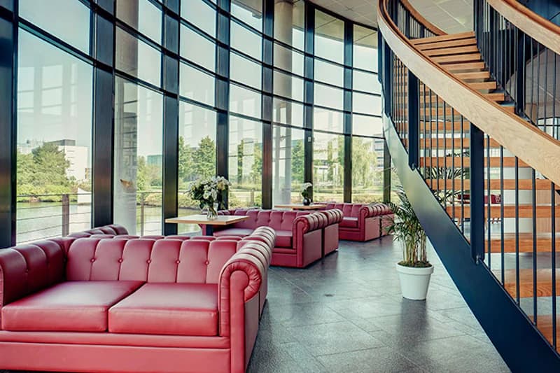 Hotellounge mit roten Ledersofas vor geschwungener Glasfront und gebogener Holztreppe.