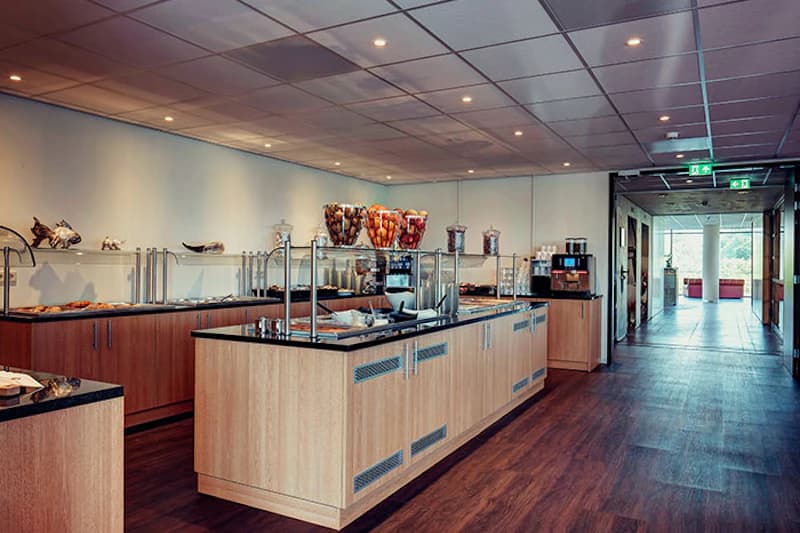 Hotelbuffet mit Holztheken, Müslispendern, Kaffeemaschine und Gang zu einem hellen Fensterbereich.