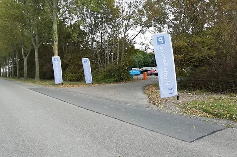 Zufahrt zu einem Parkplatz mit drei hohen weißen Fahnen, orangefarbener Schrankenpfosten und mehreren geparkten Autos hinter Büschen.
