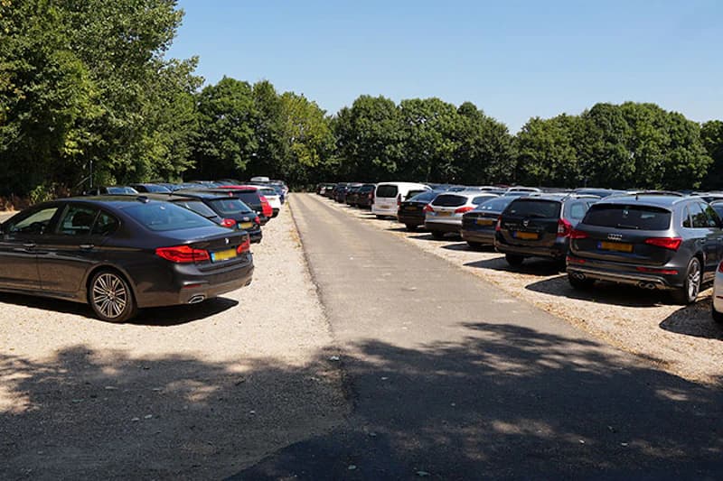Parkplatz mit zwei Reihen abgestellter Autos auf geschotterten Stellflächen und asphaltierter Mitte, Bäume im Hintergrund.