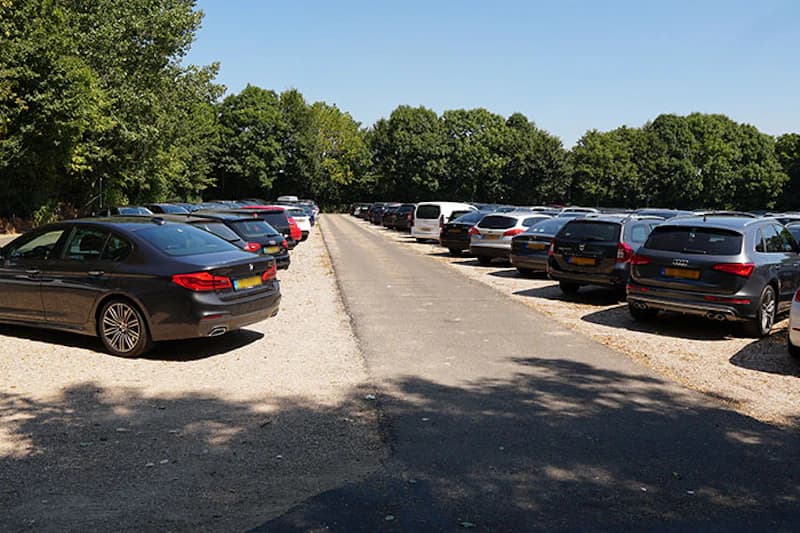 Sonniger Parkplatz mit geparkten Autos links und rechts einer zentralen Zufahrt, Bäume im Hintergrund.