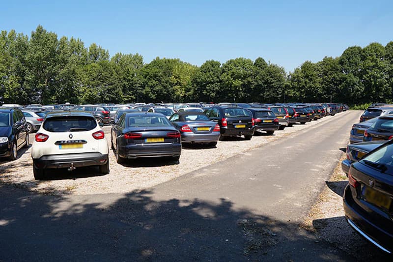 Außenparkplatz mit zahlreichen in Reihen geparkten Autos auf Schotterflächen, Fahrweg und Baumreihe im Hintergrund.