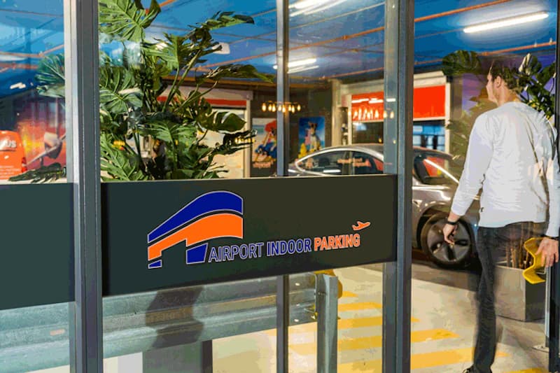 Glastür mit Airport Indoor Parking Schriftzug, Mann geht in beleuchtetes Parkhaus zu geparkten Autos.