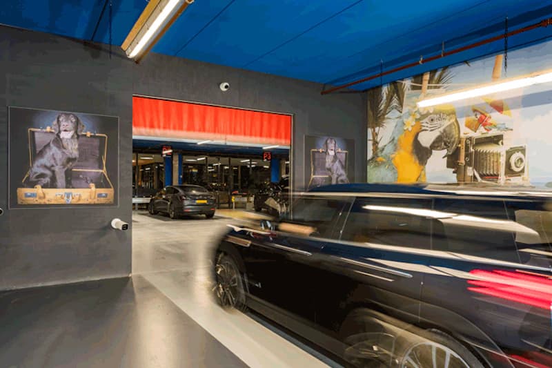 Parkhausinnenbereich am Flughafen Amsterdam mit verschwommenem einfahrendem schwarzen Auto, geparkten Fahrzeugen und bunten Wandbildern.