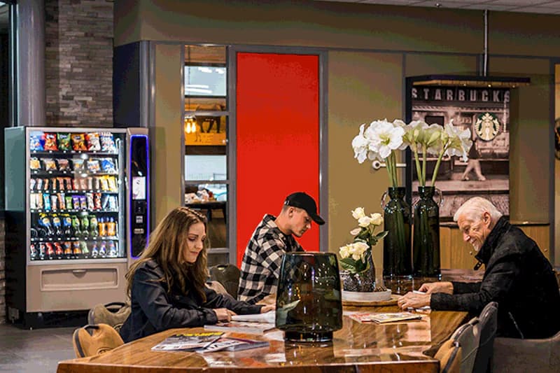 Drei Personen sitzen an langem Holztisch in einem Flughafen-Wartebereich, Snackautomat und Starbucks-Werbung im Hintergrund.