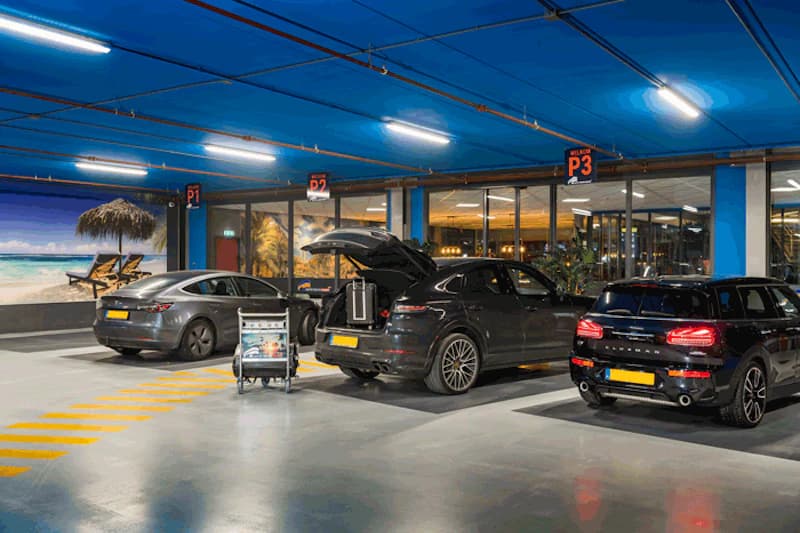 Überdachtes Parkdeck mit drei geparkten Autos, offenem Kofferraum, Gepäckwagen und Wandbild mit Strandmotiv.
