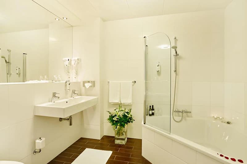 Weiß gekacheltes Badezimmer mit Doppelwaschbecken, großem Spiegel, Badewanne mit Duschabtrennung und Blumenvase.