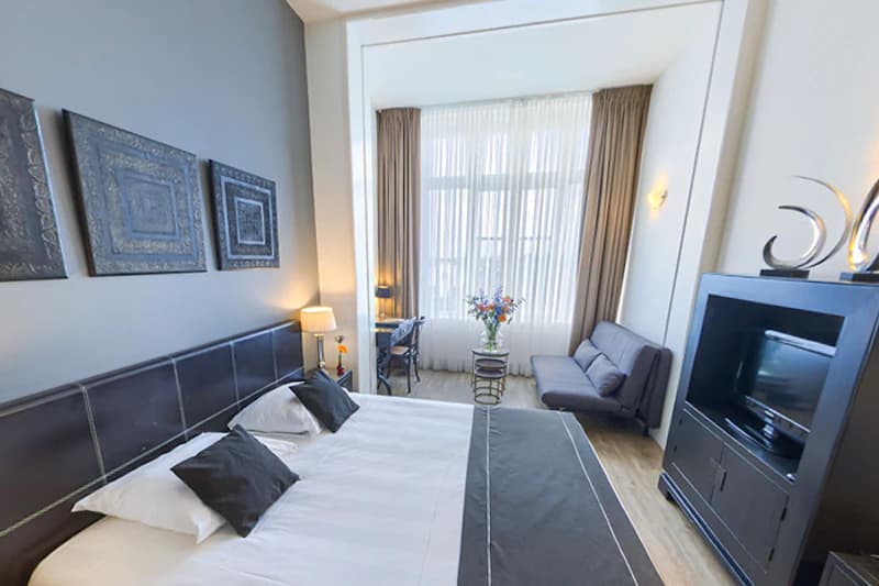 Geräumiges Hotelzimmer mit Doppelbett, Nachttischlampen, Sitzbereich, Schreibtisch und Fenster mit Vorhängen.