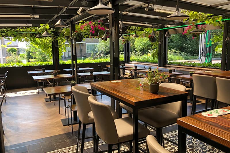 Überdachte Hotelterrasse mit Holztischen, gepolsterten Stühlen, Hängetöpfen und Blick auf eine grüne Außenanlage.