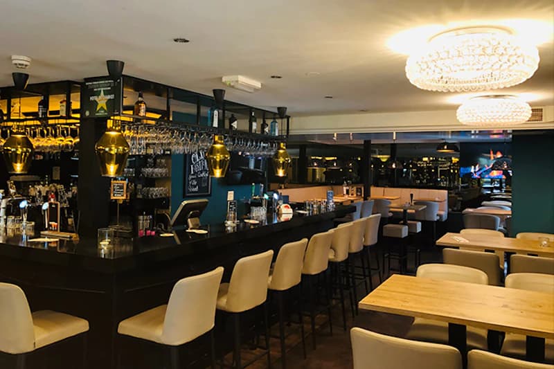 Dunkle Hotelbar mit langer schwarzen Theke, hohen Barhockern, goldenen Pendelleuchten und Tischen.