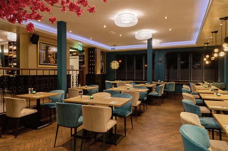 Hotelrestaurant mit mehreren Tischen, türkis- und beigen Stühlen, Kronleuchtern und Holzfußboden.