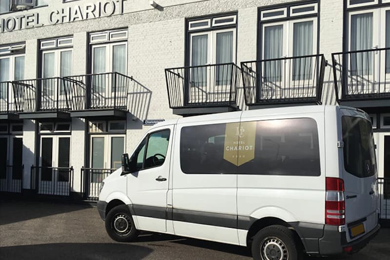 Weißer Shuttlebus mit Hotellogo vor weißem Backsteinhaus mit Balkonen und Schriftzug Hotel Chariot.