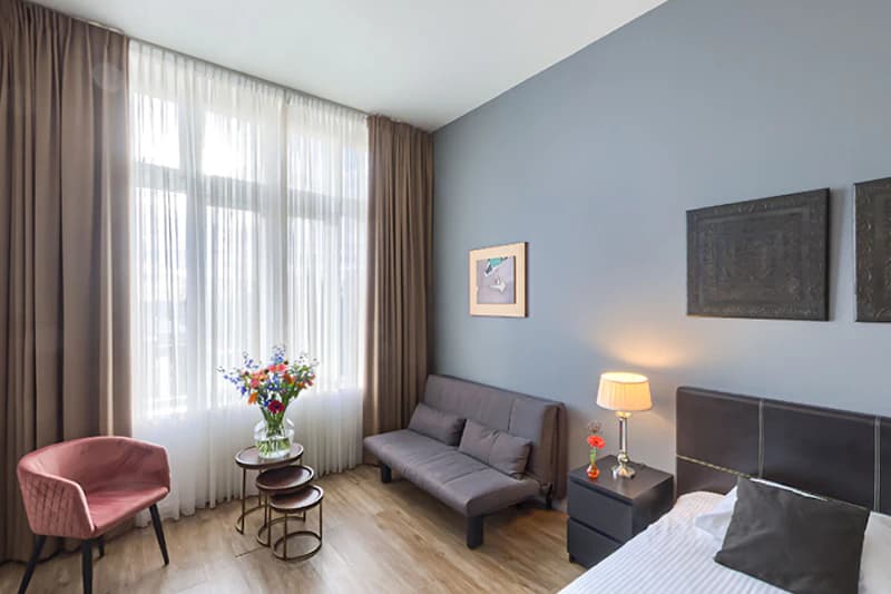 Hotelzimmer mit Bett, Sofa, Sessel, großem Fenster mit Vorhängen, Nachttisch mit Lampe und Blumenvase auf kleinen Tischen.