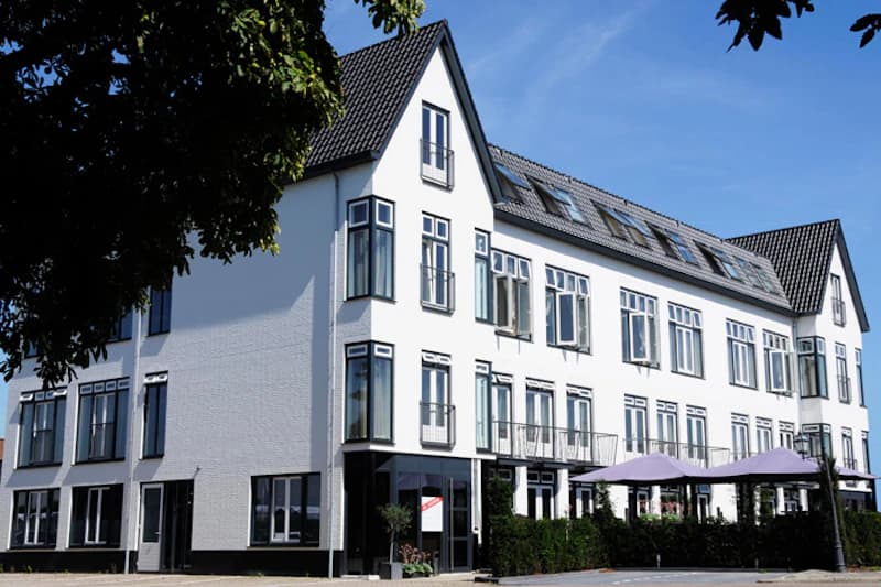 Weißes dreistöckiges Hotelgebäude mit zahlreichen Fenstern und lila Sonnenschirmen vor einer Terrasse.