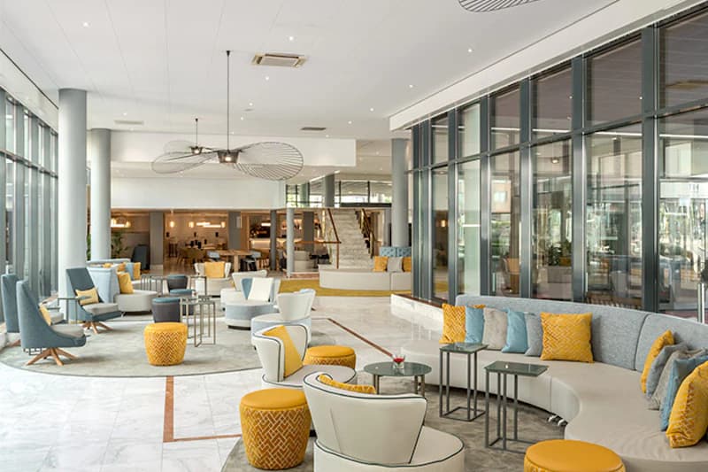 Helle Hotel-Lobby mit Sitzgruppen, gelben Kissen, großen Glasfenstern und Treppe im Hintergrund.