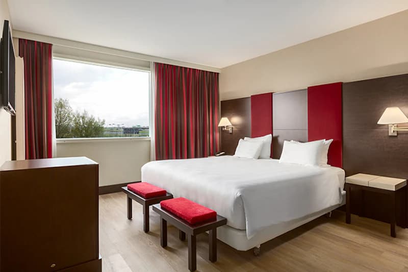 Geräumiges Hotelzimmer mit großem Doppelbett, zwei roten Hockern und Fenster mit Blick aufs Flughafengelände.