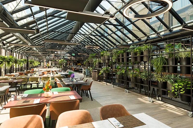 Weites Hotelrestaurant unter Glasdach mit vielen Tischen, Stühlen und Pflanzen.