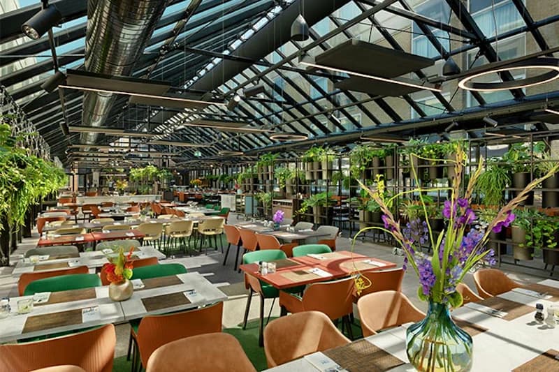 Großes helles Restaurant mit Glasdach, vielen Tischen, bunten Stühlen und Pflanzen.