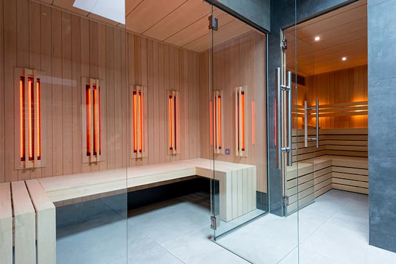 Innenraum einer Sauna mit Holzbänken, leuchtenden Heizstäben an der Wand und Glastür.