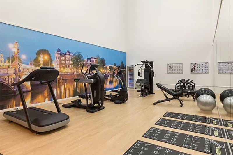 Kleines Hotel-Fitnessstudio mit Laufband, Crosstrainer, Kraftgeräten, Matten und Wandbild einer Amsterdamer Gracht.