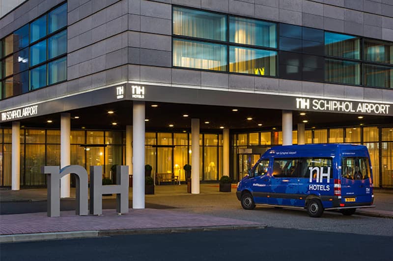 Eingangsbereich des NH Schiphol Airport Hotels mit beleuchtetem Schriftzug, großem NH-Logo und blauem Hotelbus.