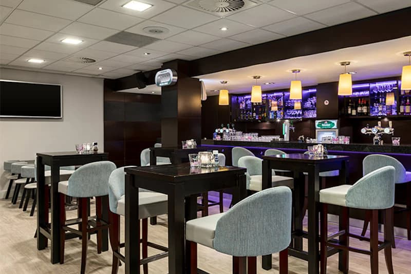 Hotelbar mit dunklem Tresen, beleuchteten Regalen, hohen Tischen und gepolsterten Barhockern.