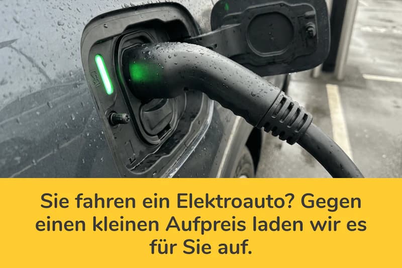 Ladestecker eines Elektroautos im Ladeanschluss mit grünem Kontrolllicht und regennasser Karosserie, darunter gelbes Banner mit Hinweistext.