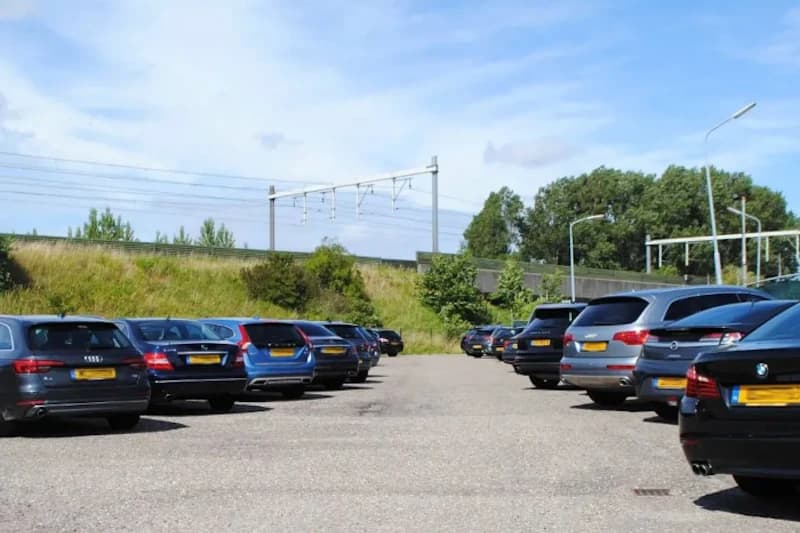 Außenparkplatz mit zwei Reihen geparkter Autos, grasbewachsene Böschung und Eisenbahnoberleitung im Hintergrund.